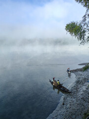 A misty morning scene over a wide river features a small wooden boat where people are preparing to fish surrounded by dense fog hanging low over the water and distant treeline an ethereal atmosphere