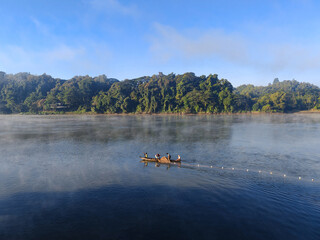 A wide shot of a tranquil river under clear blue sky captures a small wooden fishing boat carrying people navigating the calm waters, and a fishing  wall of tropical forest lines the far bank