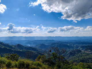 Panoramic view overlooking rolling green hills and distant blue mountain ranges under bright blue sky with dramatic white clouds covering horizon