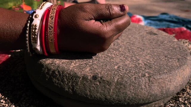 An Indian woman in a rural village uses a traditional hand-operated millstone (quern) to grind black gram lentils, showcasing authentic village life, heritage food preparation, and indigenous househol