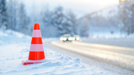 Orange traffic cone on snowy roadside blurred winter background, safety concept in cold weather conditions, road maintenance alert, icy hazard prevention, winter driving environment emergency setup