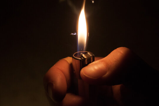 Macro close up of a classic metal lighter with a burning orange and blue flame ignited against a dark background.