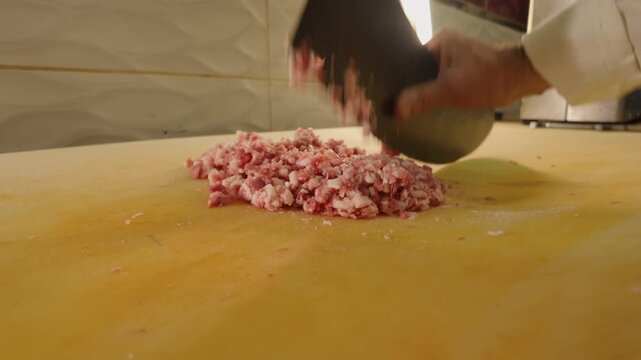 Close-up 10-bit slow motion of a chef mincing lamb meat with a traditional satir cleaver. Capturing the rhythmic process and developing meat texture for authentic Turkish kebabs. 4K quality.