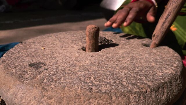 Under natural sunlight in a rural setting, an Indian woman uses a manual grinding stone to process black gram lentils, reflecting cultural traditions and self-reliant domestic practices.