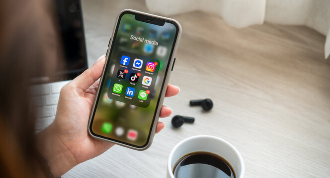 Bangkkok, THAILAND - January 2nd, 2026: hands of woman Iphone with icons of social media on screen place on table with tablet and ear phone and a mug of coffee
