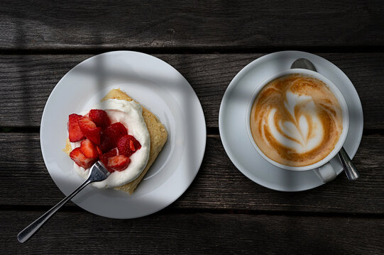 A strawberry biscuit and a cup of cappuccino are on a wooden table.