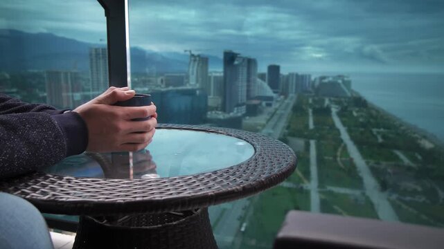 man drinking tea on balcony Batumi skyline and Black Sea view at dusk, hands holding ceramic cup on wicker table, distant promenade and highrises fading into mist, solitary traveler reflecting