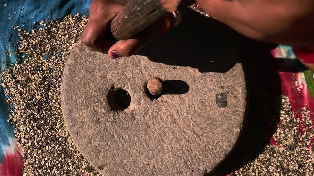 Under natural sunlight in a rural setting, an Indian woman uses a manual grinding stone to process black gram lentils, reflecting cultural traditions and self-reliant domestic practices.