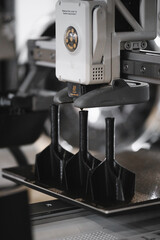Industrial 3D printer extruder head printing multiple black plastic parts on build plate during additive manufacturing process. Close-up of heated 3D printer nozzle