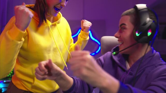 Two woman gamers wearing headsets playing a video game together in a colorful gaming room. They are holding game controllers, interacting with the screen, and enjoying an exciting multiplayer session