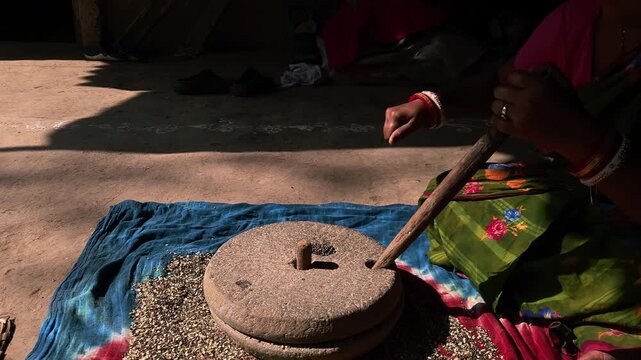 Under natural sunlight in a rural setting, an Indian woman uses a manual grinding stone to process black gram lentils, reflecting cultural traditions and self-reliant domestic practices.