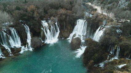 Aerial horizontal view of Kravica Waterfalls in Bosnia during winter, with powerful cascades flowing into a turquoise pool, surrounded by bare trees and rocky cliffs in a serene natural landscape © Ekaterina