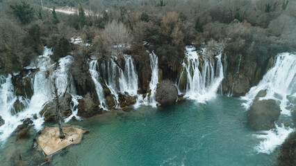 Aerial horizontal view of Kravica Waterfalls in Bosnia during winter, with powerful cascades flowing into a turquoise pool, surrounded by bare trees and rocky cliffs in a serene natural landscape © Ekaterina