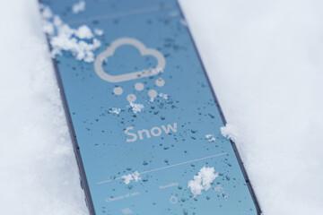 Smartphone lying in snow outdoors with weather app screen showing snow forecast