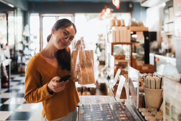 Asian woman shopping at local coffee shop, holding takeaway order while paying with smartphone. Authentic lifestyle, small business storytelling, and consumer experience content.