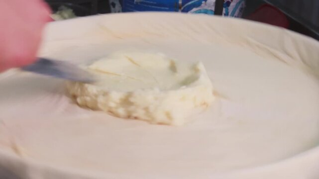 Close-up 10-bit slow motion of an artisan spreading traditional semolina cream (kaymak) over thin baklava dough. A key step in preparing authentic Gaziantep baklava in a professional bakery.