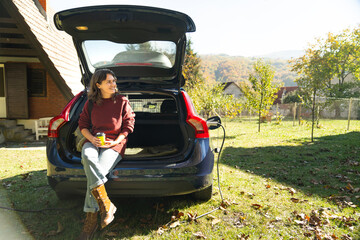 Woman traveler with backpack sitts into trunk of charging electric car