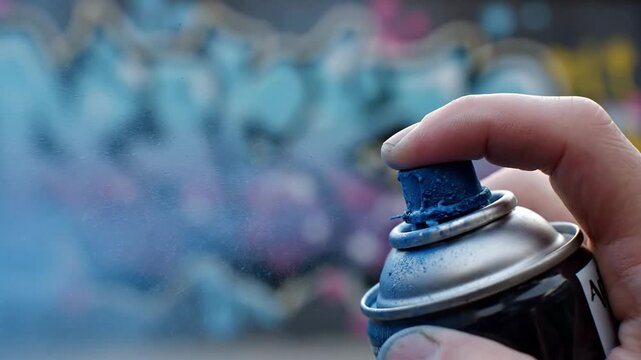 Close up of a hand holding a spray paint can spraying blue paint onto a vibrant graffiti wall with purple and teal colors in the background outdoor daytime lighting