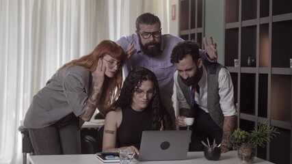 A diverse group of creative professionals collaborates while looking at a laptop screen in a modern office setting.