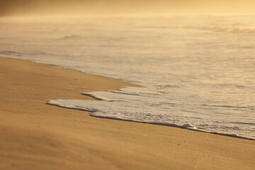 Detalhe da mar&eacute; avan&ccedil;ando suavemente sobre a areia da praia de Itamambuca ao amanhecer, com luz dourada e atmosfera tranquila.