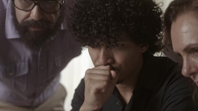 Diverse group of people looking seriously at a screen with focus on a pensive young man with curly hair.