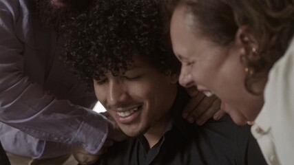 Young man with curly hair smiling with eyes closed while friends place hands on his shoulders for support and comfort during a group gathering.