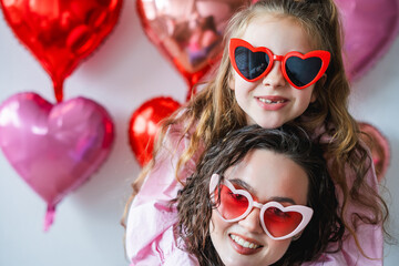 Cute little girl with her teenage sister in heart glasses on a background of pink balloons. Valentine's Day.