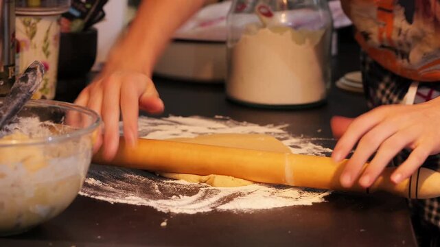 Short handheld close-up video in a home kitchen shows eclair dough being rolled with a wooden rolling pin on a black textured countertop, with utensils and bowls nearby, 2025