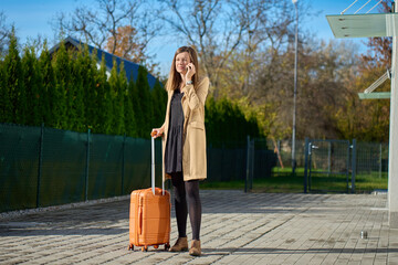 Woman in beige coat talking on phone and holding orange suitcase outside