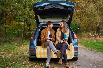 Man and woman sitting on open car trunk with plaid and thermos cups