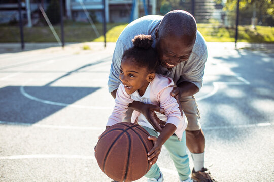 Senior grandfather and child granddaughter laughing while playing basketball on outdoor court