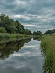 Obraz premium Overcast day reflecting dark forest and grassy banks on calm slow moving river water beneath dramatic gray clouds