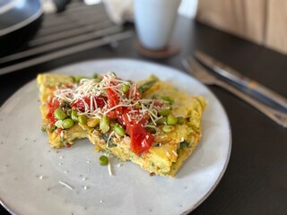 Vegetable omelette slice with tomatoes, peas and cheese on plate; a homemade vegetable omelette served on a ceramic plate, topped with tomatoes, green peas and grated cheese