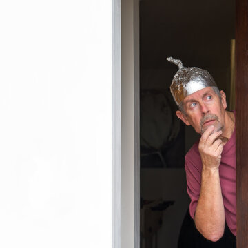 Tinfoil hat picture. A man wearing a tinfoil hat looks anxiously out of a door towards the sky. Paranoia and conspiracy theory. Alien abduction and government cover up. Mind control and brainwashing. 