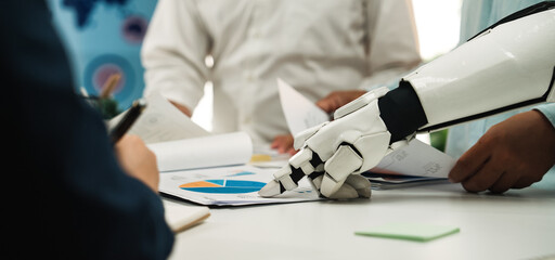 A futuristic robot arm points to business data and charts during a collaborative meeting, showcasing the integration of technology in modern corporate strategy discussions.Noogenesis