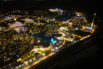 Fototapeta premium Nighttime Aerial of the Faro de Maspalomas Lighthouse, El Oasis Palm Grove, and the Elite 5-Star Resort Strip of Meloneras, Gran Canaria, Spain