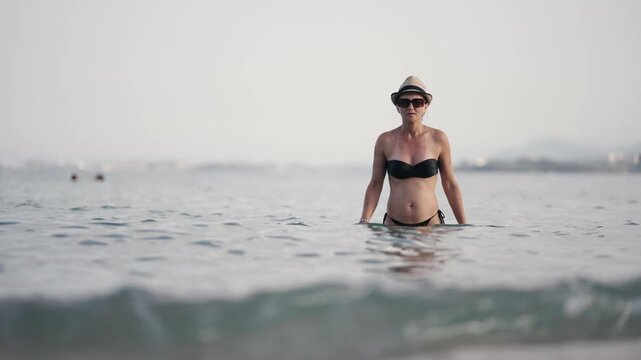 Attractive woman wearing nat and bikini relaxing walk in calm sea water deep up to waist as water splashing her fit body. Enjoying summer vacation concept, slow motion
