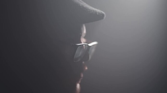 Dark moody side portrait of caucasian man with cap and beard wearing glasses, cinematic shot with selective focus