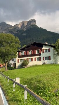 Sufers, Switzerland. Historic village church and chalets in a sunny, green Alpine meadow. The view includes a dramatic mountain panorama of rugged peaks under heavy storm clouds.