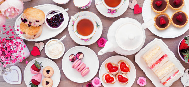 Valentines Day or love theme afternoon tea. Overhead view table scene against a wood banner background. Assortment of finger sandwiches, sweet desserts and pastries.