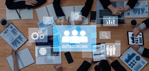 A diverse team collaborates in a business meeting focused on outsourcing strategies, utilizing digital devices and analyzing data for effective decision-making. Trope