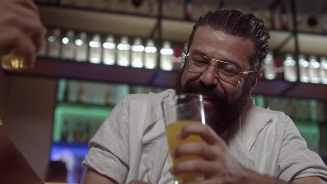 Happy middle-aged man with beard and glasses enjoying a drink at a bar during a social gathering with friends.