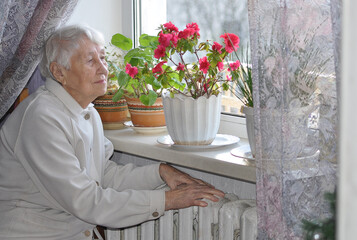 Old lonely woman sitting near the window in his house.