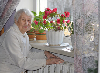 Old lonely woman sitting near the window in his house.