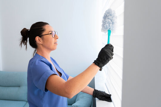 Professional cleaning woman wearing gloves and uniform diligently dusting window blinds in a bright home or office setting, ensuring a spotless and hygienic environment
