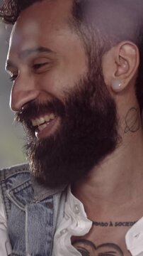 Close-up portrait of a smiling bearded man with a trendy haircut and ear plug in a dimly lit indoor setting.