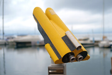 Coin operated yellow binocular viewer on blurred background of pier with boats, selective focus