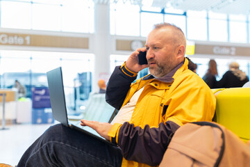 Business traveler talking on smartphone while using laptop at airport gate. Modern multitasking, communication and remote work during travel lifestyle in terminal