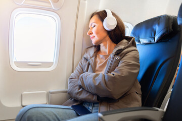 Adult caucasian woman resting with eyes closed wearing headphones inside airplane seat. Calm travel...
