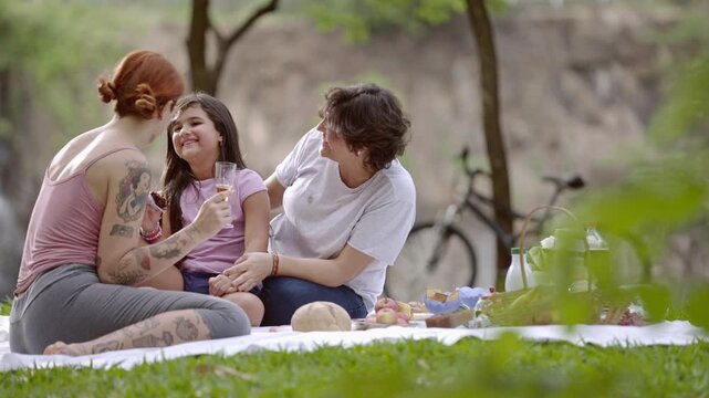 Happy lesbian couple with daughter enjoying a picnic in the park. Diverse family with tattooed mother spending quality time outdoors on a sunny day.